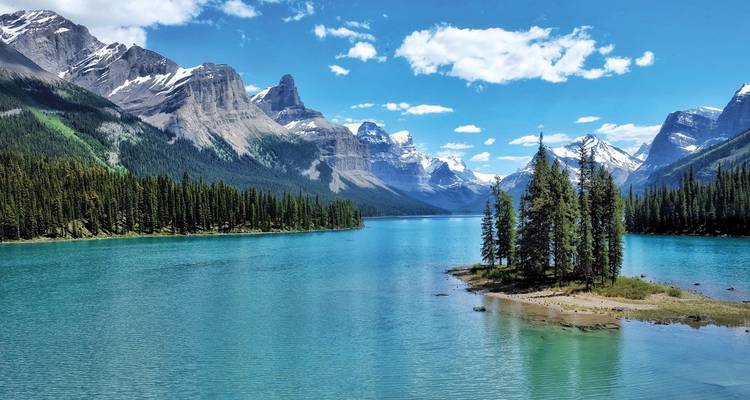 Crystal-blue lake fringed by evergreen forest and dramatic Rocky Mountain peaks under a bright sky.