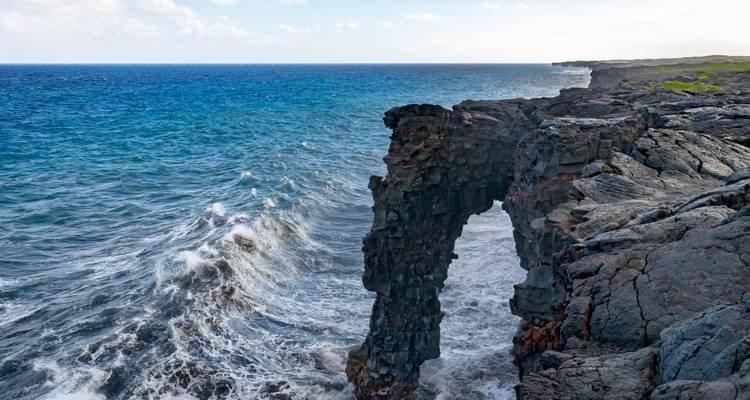 Dramatic volcanic sea cliff forming a natural stone arch with waves crashing below and a vast blue ocean horizon