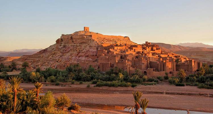 Die irdene Ksar von Ait Benhaddou liegt auf einem Hügel, der in warmes Abendlicht getaucht ist, über einem trockenen Flussbett.