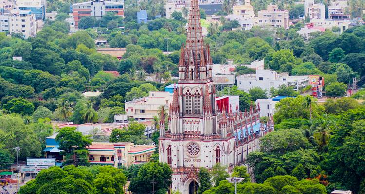 Une haute flèche d'église néo-gothique s'élève au-dessus d'arbres verts luxuriants et d'un paysage urbain bas, se détachant dans le centre de Trichy.