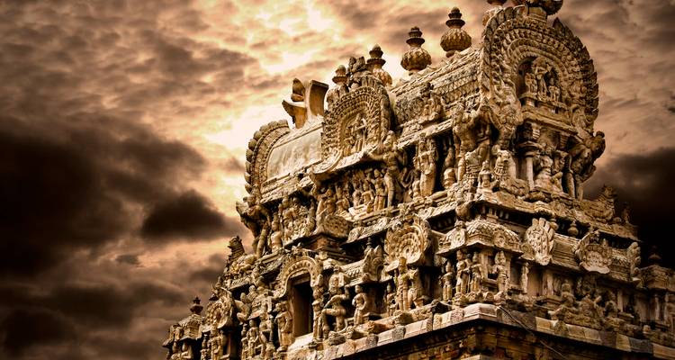 Une tour de temple en pierre patinée riche en sculptures est dramatiquement éclairée sous un ciel maussade aux teintes bronze.