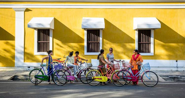 Six femmes en saris colorés discutent à côté de vélos colorés devant un mur colonial jaune vif à Puducherry.