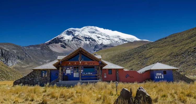 Berghütten-Gebäude auf grasbewachsenen Ebenen mit dem schneebedeckten Vulkan Chimborazo, der dahinter aufragt