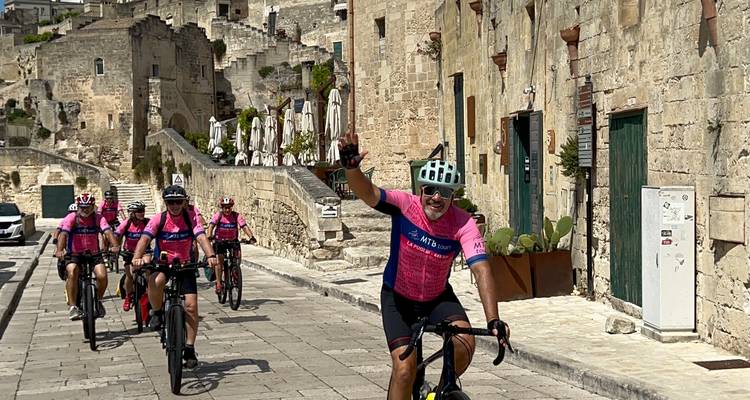 Des cyclistes roulent dans les rues étroites en pierre de Matera, l'un d'eux faisant signe à la caméra.