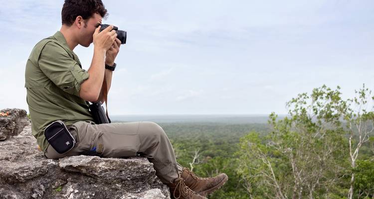 Jonge man zittend op een rotsige uitstulping die foto's maakt van een eindeloos groen bos dat zich uitstrekt tot de horizon onder een heldere hemel