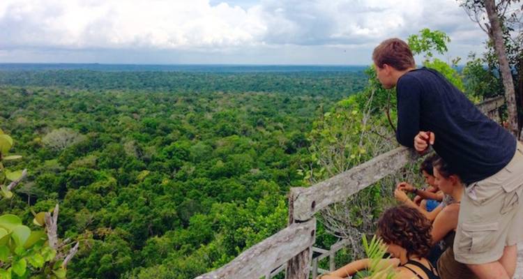 Kleine groep reizigers leunend over een rustieke houten reling, uitkijkend over een uitgestrekt jungle bladerdak onder een gedeeltelijk bewolkte hemel