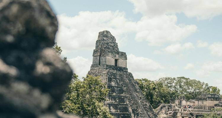 Stone Maya pyramid temple rising above the surrounding trees with soft clouds in a blue sky