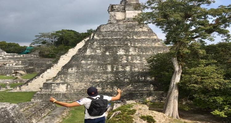 Traveler with backpack raising both thumbs while facing a massive stepped Maya pyramid in lush jungle surroundings