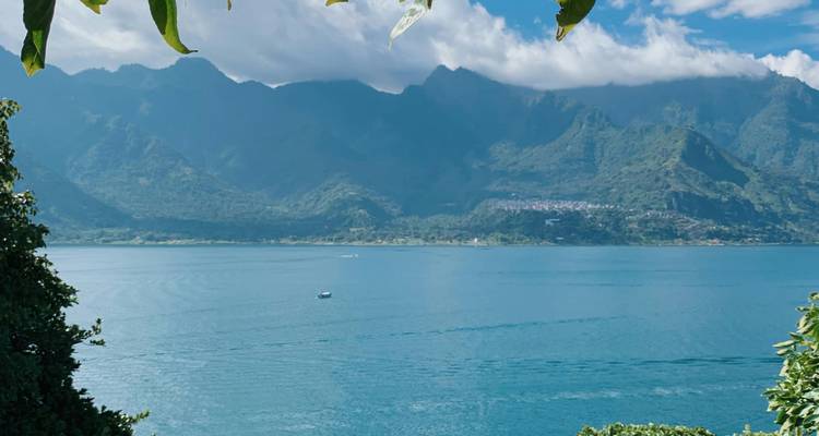 Deep blue lake bordered by dramatic green mountains under scattered clouds, framed by leafy branches