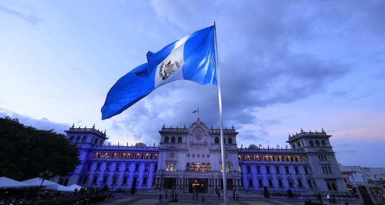 Large Guatemalan flag waving before an illuminated neoclassical government palace at dusk
