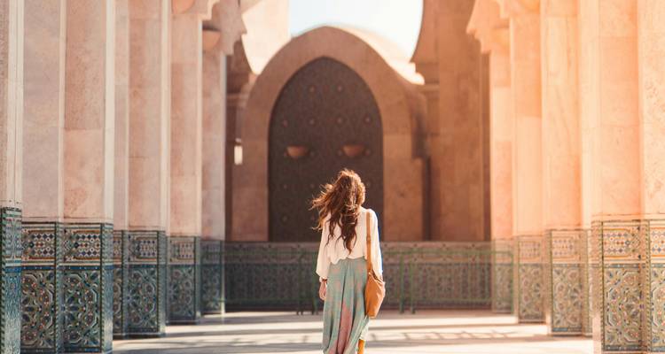 Vista posterior de una mujer caminando a través de la arcada de mármol ornamentada de la Mezquita Hassan II bañada en luz cálida.