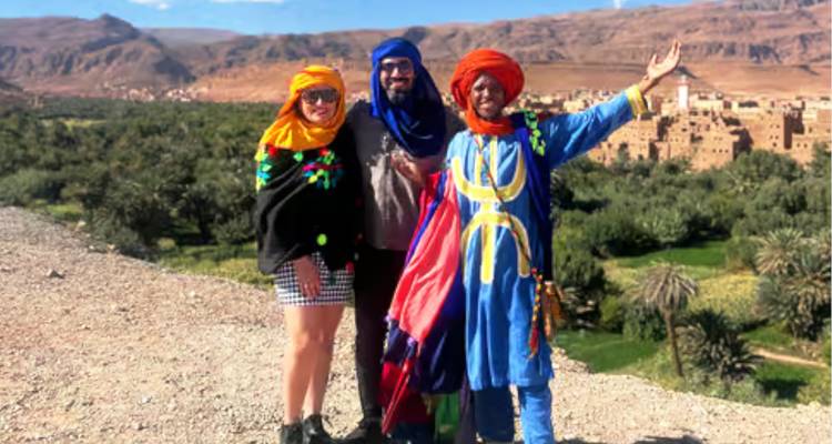 Trío de turistas posando con guía bereber en túnicas coloridas con vista a un valle exuberante.