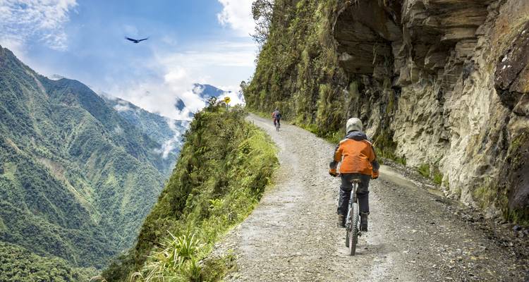 Des cyclistes roulent sur une route de gravier étroite accrochée au flanc escarpé d'une montagne verdoyante avec de la brume et un oiseau qui plane au-dessus