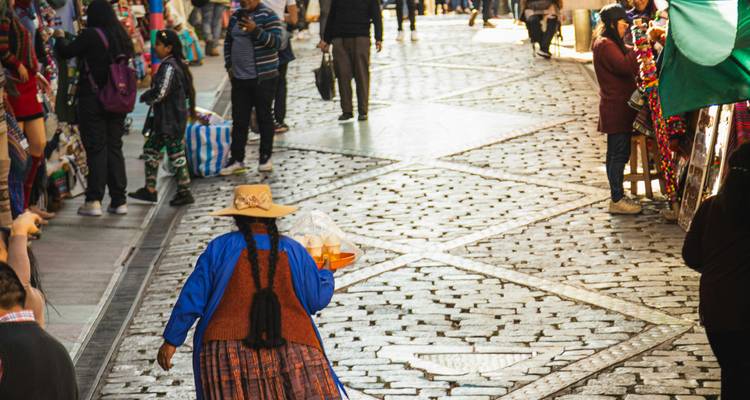 Marché de rue animé aux pavés avec une femme vêtue traditionnellement portant un plateau et des acheteurs parcourant des étals colorés