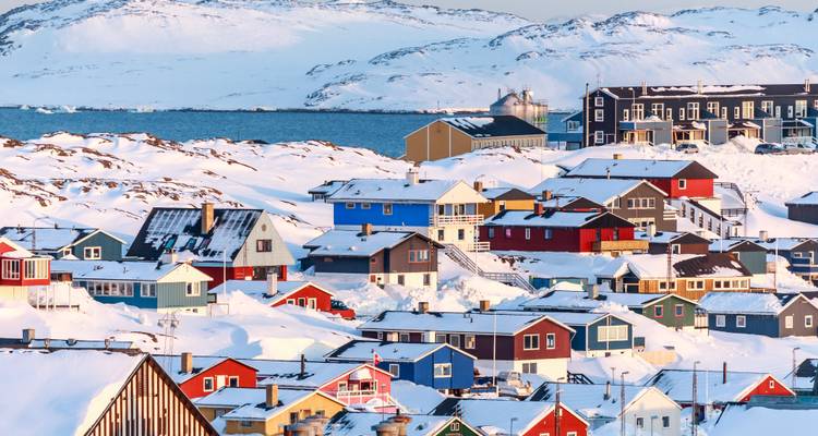 Maisons pastel couvertes de neige d'un village du Groenland au bord d'une mer bleue glacée et de collines blanches.