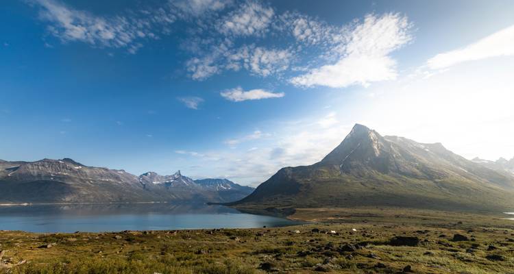 Large fjord groenlandais avec des montagnes escarpées, une eau calme et une toundra ouverte sous un ciel lumineux.