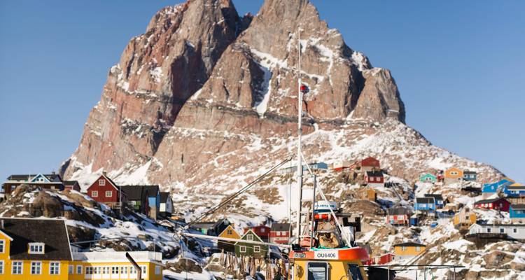Pic de granit acéré s'élevant au-dessus d'un groupe de maisons colorées saupoudrées de neige et d'un port en activité au Groenland.