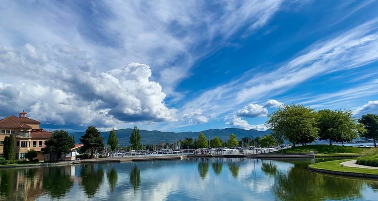 Reflective pond mirrors dramatic clouds and distant mountains beside a lakeside promenade.