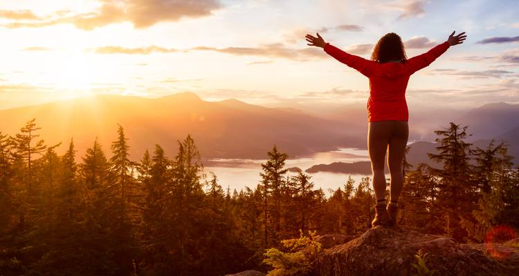Hiker standing triumphant on a rocky outcrop overlooking forested mountains and shining water at sunrise