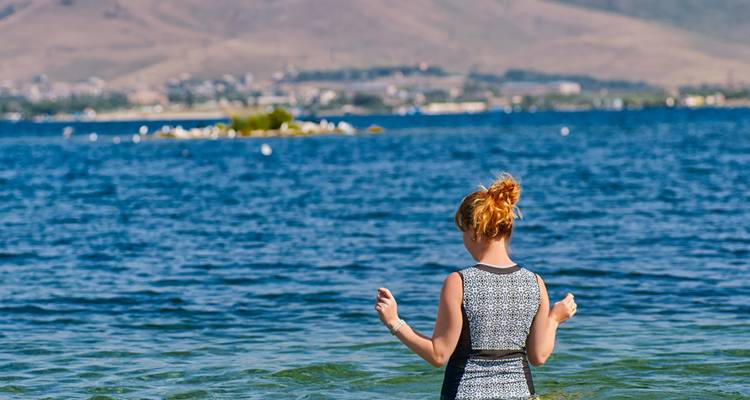 Une femme en robe à motifs s'avance dans les eaux bleu profond du lac Sevan avec des collines au loin derrière elle.