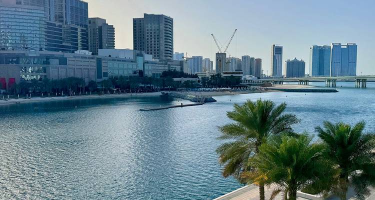 Waterfront promenade lined with palm trees overlooks calm blue water and high-rise buildings of Abu Dhabi.