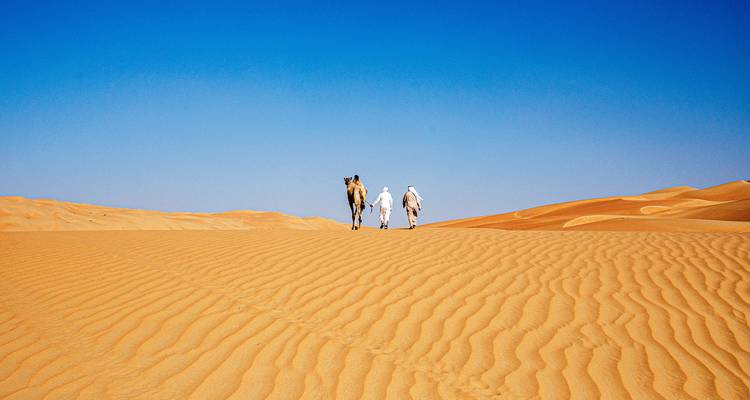 Two robed travellers and a camel walk across vast golden sand dunes beneath a cloudless desert sky.