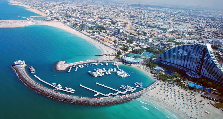 Aerial view of Dubai’s curved marina, jetties and turquoise waters framed by sandy coastline and the distinctive wave-shaped Jumeirah Beach Hotel.