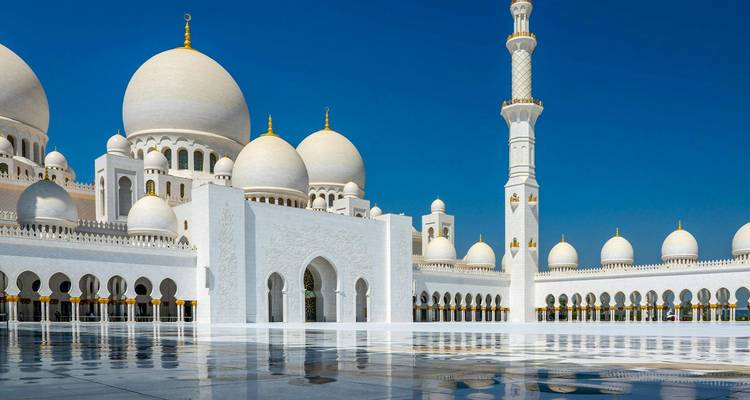 Witte marmeren moskee met meerdere koepels en een hoge minaret die zich weerspiegelt op een glanzende binnenplaats onder een helder blauwe lucht.