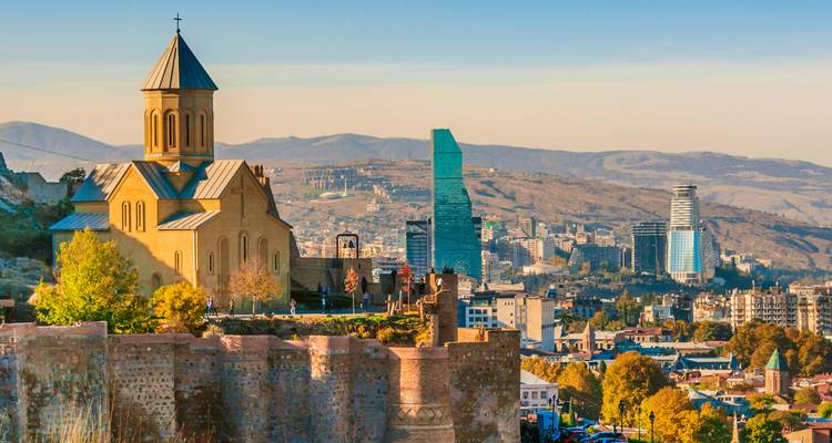 Une église dorée et d'anciens murs de forteresse dominent les tours modernes dans une vue panoramique de Tbilissi au coucher du soleil.