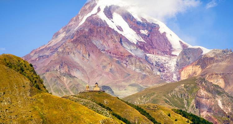 Le majestueux mont Kazbek s'élève au-dessus de collines verdoyantes vallonnées avec l'église de la Trinité de Gergeti visible sur une crête lointaine.