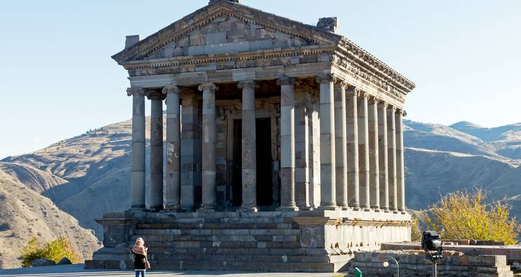 Der klassische säulengeschmückte Tempel von Garni steht auf einem Felsvorsprung mit Blick auf die zerklüfteten armenischen Hügel mit einem einzelnen Besucher im Vorhof.