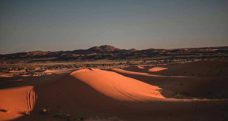 Dunas desérticas besadas por el sol brillando en naranja con colinas rocosas distantes bajo un cielo vespertino que se oscurece.