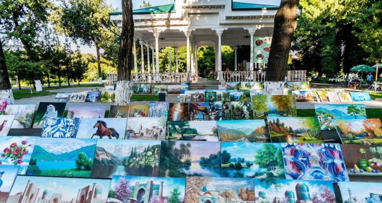 Marché d'art en plein air présentant de nombreuses peintures paysagères colorées dans un cadre de parc ombragé.
