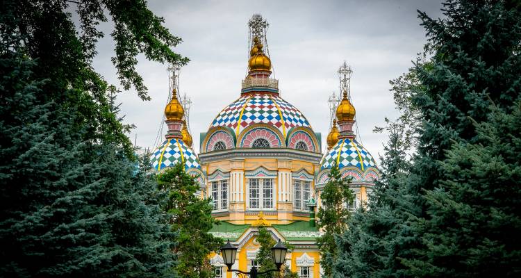 Kleurrijke koepelvormige Russisch-orthodoxe kathedraal omlijst door hoge naaldbomen tegen een bewolkte hemel.