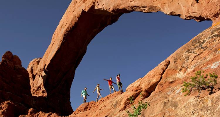 Des randonneurs marchant sous une immense arche de grès rouge massif sur un ciel bleu profond