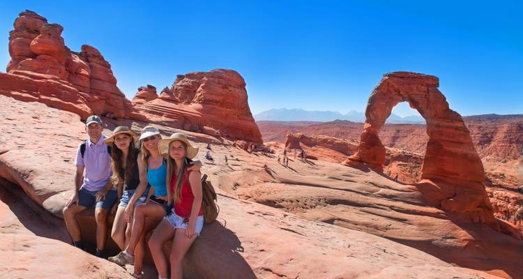 Les visiteurs sont assis sur une corniche de grès rouge face à l'emblématique Arche Délicate sous un ciel bleu profond