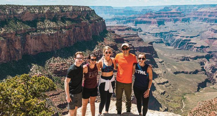 Los amigos están del brazo en un mirador del Gran Cañón con vastos acantilados detrás de ellos.