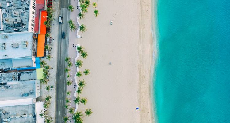 Bovenaanzicht vanuit drone van zandstrand, turquoise zee en gebouwen aan het strand omzoomd met palmbomen.