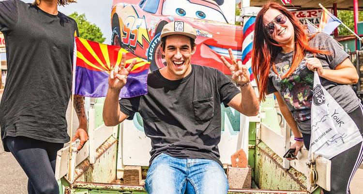 Smiling road-trippers posing with Arizona flag and vintage truck at roadside attraction