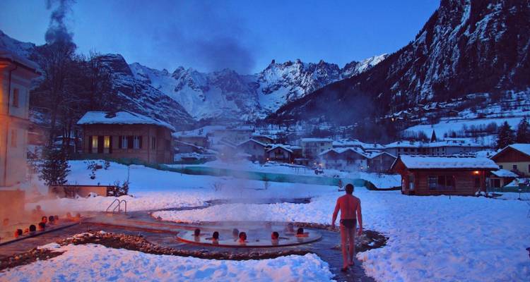 Bather walking toward steaming outdoor hot spring pool amid snowy alpine village at dusk.
