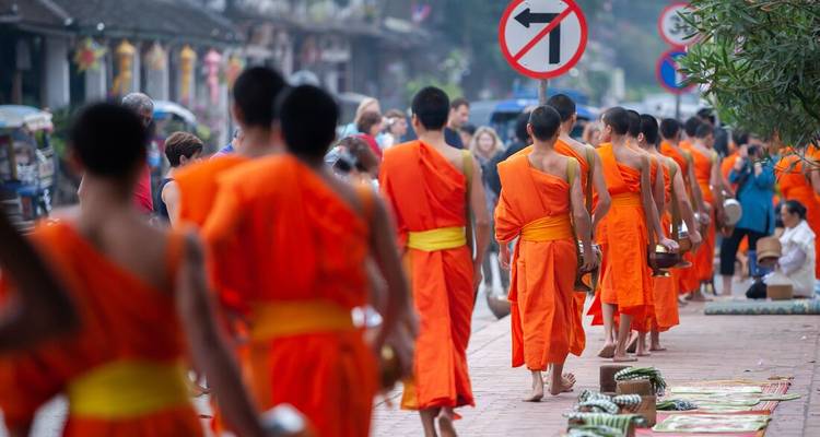 A long line of saffron-robed Buddhist monks walk barefoot along a city sidewalk during alms collection.