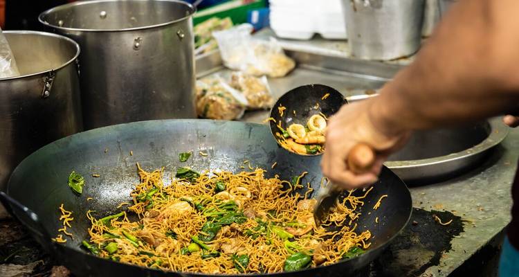 Street-food chef stir-fries noodles with vegetables in a black wok at an open kitchen stall.