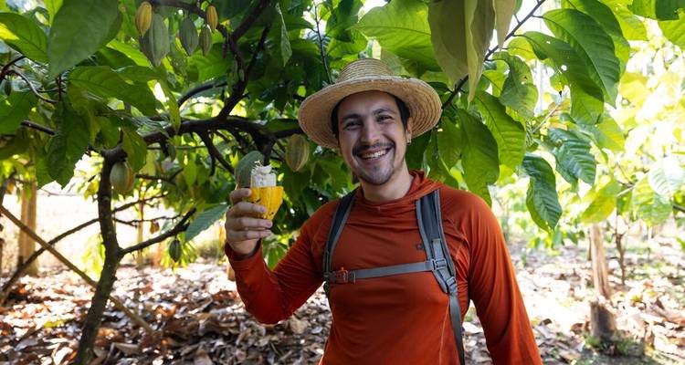 Smiling traveler wearing a straw hat holds fresh cacao fruit under lush green trees.