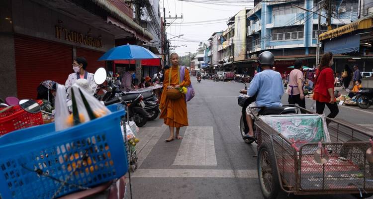 Urban street scene featuring a lone monk with an alms bowl amid scooters and market traffic.
