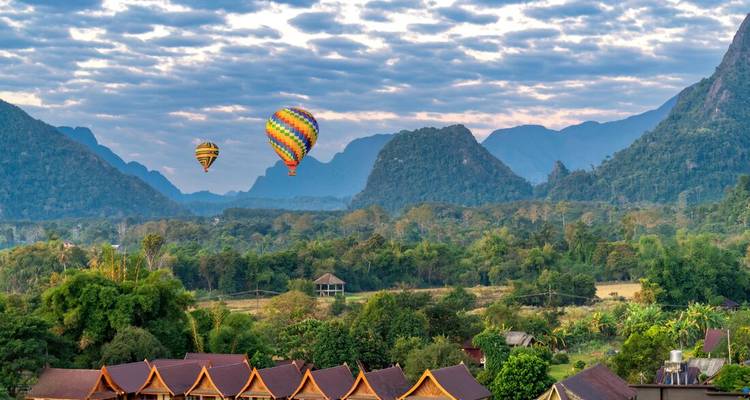 Colorful hot-air balloons rise over lush valleys and jagged limestone mountains at sunrise.