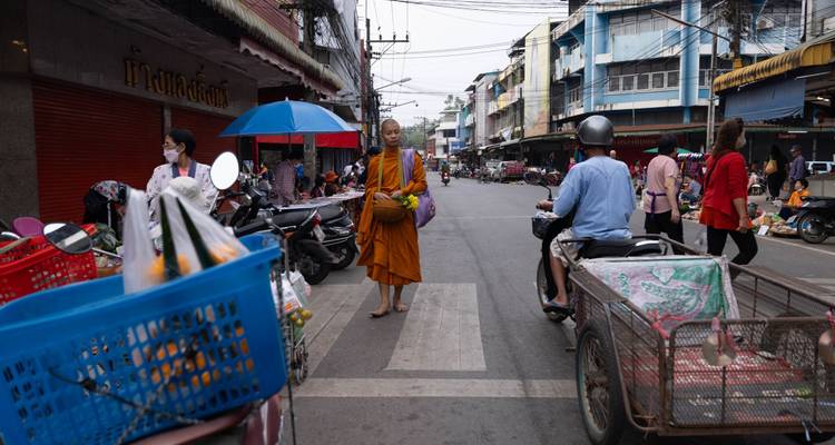 Buddhistischer Mönch, der Almosen in einem belebten südostasiatischen Straßenmarkt mit Rollern und Verkäufern sammelt.