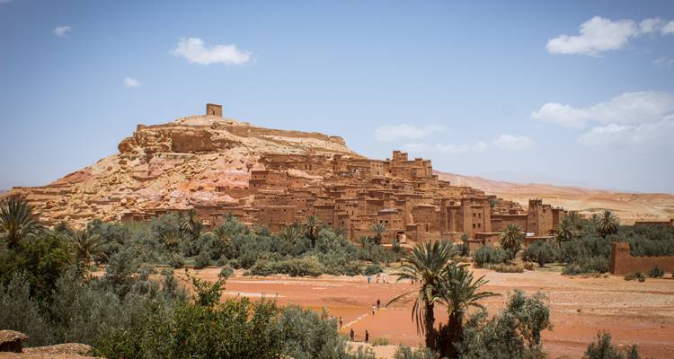 The earthen ksar of Ait Ben Haddou rises above a verdant river oasis under a blue sky.