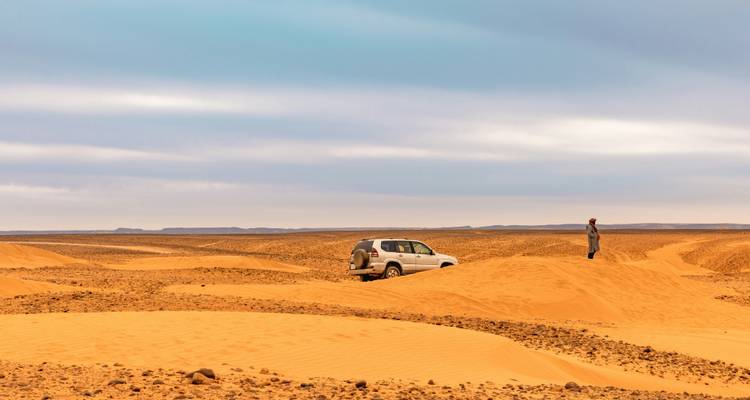 SUV aislado y viajero entre dunas de arena ondulantes bajo un vasto cielo desértico nublado.