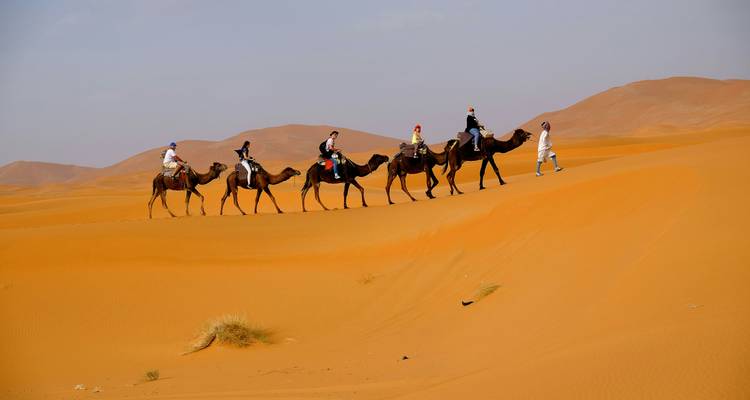Fila de turistas montando camellos guiados por un guía a través de dunas de desierto anaranjadas bajo un cielo brumoso.
