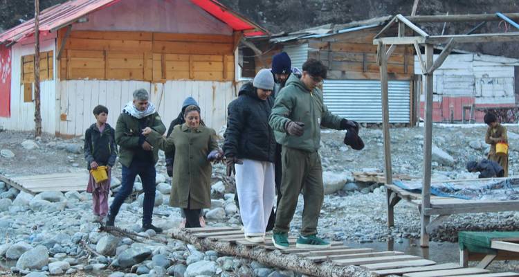 Des voyageurs traversent prudemment un étroit pont de planches en bois au-dessus d'un lit de rivière rocailleux.
